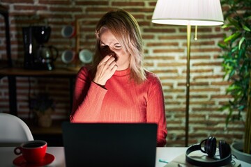 Canvas Print - Blonde woman using laptop at night at home smelling something stinky and disgusting, intolerable smell, holding breath with fingers on nose. bad smell