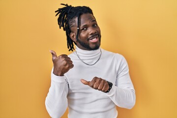 Poster - African man with dreadlocks wearing turtleneck sweater over yellow background pointing to the back behind with hand and thumbs up, smiling confident