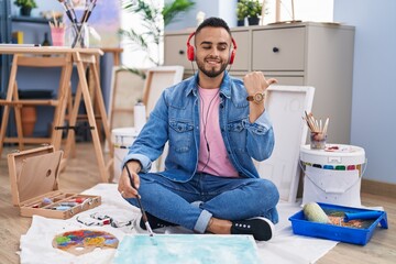 Poster - Young hispanic man painter sitting on the floor at art studio pointing thumb up to the side smiling happy with open mouth