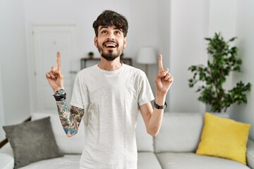 Poster - Hispanic man with beard at the living room at home smiling amazed and surprised and pointing up with fingers and raised arms.