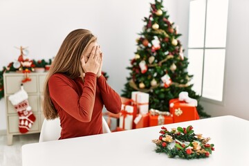 Canvas Print - Caucasian young blonde woman sitting on the table by christmas tree with sad expression covering face with hands while crying. depression concept.