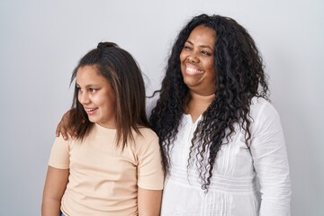 Poster - Mother and young daughter standing over white background looking away to side with smile on face, natural expression. laughing confident.