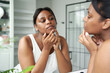 © gpointstudio - Young African-American woman squeezing a pimple in the bathroom mirror