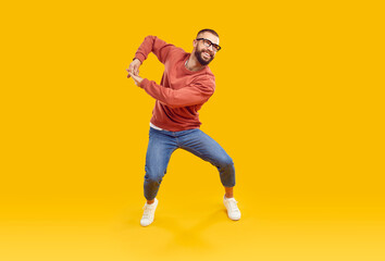 Poster - Cheerful male dancer or fashion model in casual clothes dancing and having fun in studio. Happy handsome young man in orange sweatshirt, blue jeans and glasses dancing isolated on yellow background