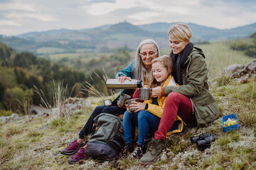 Small girl with mother and grandmother sitting and drinking hot tea on top of mountain in autumn day.