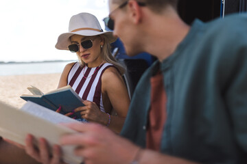 Wall Mural - Young couple sitting together in front of van, camping and reading book.