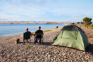 Wall Mural - A married couple with a dog are resting on the shore of a lake in a camping. Family vacation with a tent in nature.