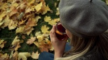 Close Up Of Woman Sits On Autumn Maple Leaves And Drinks Hot Mulled Wine With Anise And Lemon In Park Outdoors. Woman With Tea On Lawn In Garden