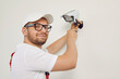 © Studio Romantic - Portrait of smiling male technician worker who installing video surveillance camera. Young man in cap and glasses, who fastens surveillance camera to white wall with screwdriver, looks into camera.