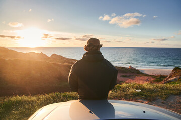 Back view of the young man looking at the sea and relaxing while sitting at his car