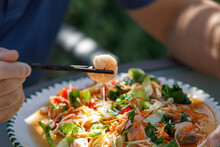 Chopsticks Hold A Prawn From A Homemade Seafood Papaya Salad With Prawns And Salmon On A White Plate With Male Hands And Chopsticks, Outdoors In Sun And Shade