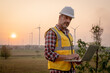 © Jirus - Portrait of engineer wearing yellow vest and white helmet using a computer laptop on site at wind turbines field or farm, Sustainable energy concept
