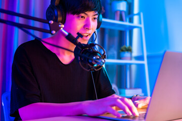 Wall Mural -  Young asian handsome man sitting on chair holding cellphone in his hand. Exited streamer wearing headset in online mobile game in neon room .Esport streaming game online