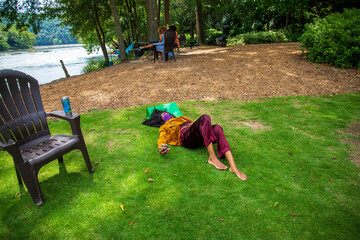 an African American woman wearing a brown jacket and burgundy pants and a mask laying on the lush green grass in a gorgeous summer landscape on the Chattahoochee river surrounded lush green trees