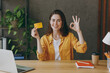 © ViDi Studio - Young successful employee business woman 20s in casual yellow shirt hold in hand mock up of credit bank card show ok gesture sit work at wooden office desk with pc laptop. Achievement career concept
