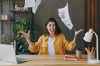 © ViDi Studio - Young cheerful fun happy successful employee business woman 20s she wear casual yellow shirt throw up paper account documents sit work at wooden office desk with pc laptop. Achievement career concept.