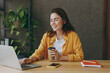 © ViDi Studio - Young happy successful employee business woman wear casual yellow shirt hold brown cup coffee hold use on laptop pc computer sit work at wooden office desk with pc laptop. Achievement career concept.