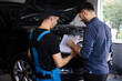 © uflypro - Manager Checks Data on a Tablet Computer and Explains the Breakdown to Mechanic. Car Service Employees Inspect the Bottom and Skid Plates of the Car. Specialist is Showing Info on Tablet.