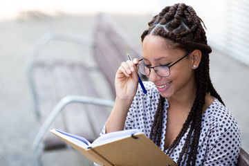 Wall Mural - Smiling young african woman writing a book sitting outside