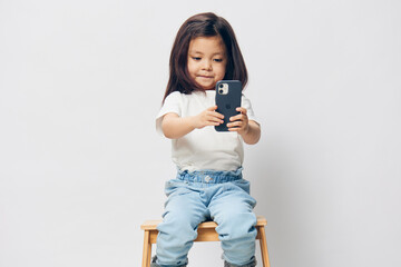 Wall Mural - a cute little girl of preschool age is standing on a white background in a white T-shirt, thoughtfully sitting on a chair and looking at the phone. The theme of children's happiness