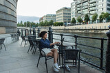 Fototapeta Uliczki - full length of young freelancer using laptop while sitting on summer terrace near river in berlin.