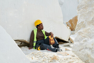 Poster - Young restful female worker in uniform and safety helmet having break after work while sitting against huge marble rock at quarry