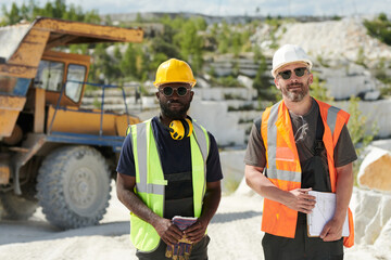 Aufkleber - Two multicultural male workers of marble quarry standing in front of camera against dump truck on territory of industrial factory