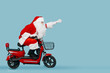 Full length side view photo of a santaclaus who rides a red electric moped with his arm extended forward in a superman pose on a blue isolated background with copy space