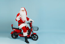 Full Length Santa Claus Sitting Behind The Wheel Of A Red Electric Moped Makes A Horn Gesture With His Hand And Looks At The Camera On A Blue Isolated Background With Copy Space
