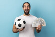Portrait of man with beard wearing white T-shirt holding soccer ball and lots dollar banknotes, looking camera with amazed face, betting and winning. Indoor studio shot isolated on blue background.