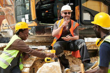 Group of happy middle aged intercultural workers of marble quarry having lunch and communicating while sitting by excavator