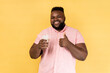 Portrait of satisfied smiling happy man wearing pink shirt feels thirsty, holding glass of cold fresh water with ice, showing thumb up. Indoor studio shot isolated on yellow background.