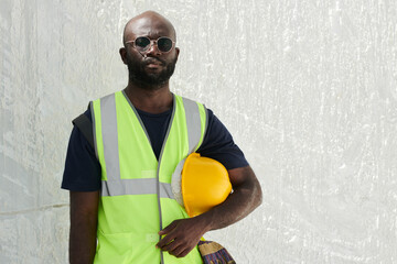 Aufkleber - Serious black man in reflective vest and eyeglasses holding hardhat by his waist while standing in front of camera against white wall