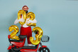 Young adult hipster man in a santa costume and big heart-shaped glasses holds inflatable numbers 2023 in his hands on a red electro scooter on a blue isolated background