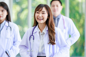 Closeup shot of Asian young cheerful happy professional intern practitioner doctor in white lab coat with stethoscope standing smiling in front of male and female colleagues on blurred background