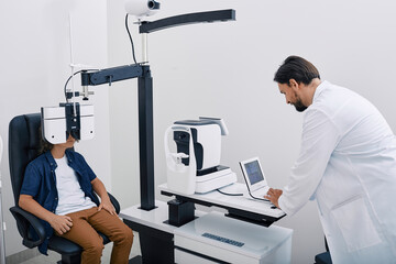 Wall Mural - Optometrist doing vision test for male child using modern phoropter at ophthalmology clinic. Eye exam and vision diagnostic in children