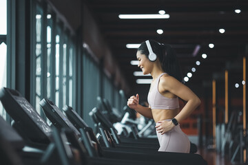 Side view of beautiful young asian woman running on treadmill and listening to music via headphone during sports training in a gym.