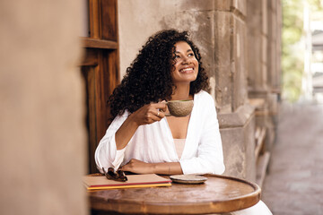 cheerful young african woman with curly brunette hair enjoying cup of coffee in spring morning. brun