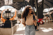Stylish young african woman walks through railway station with smartphone and cup of coffee. Girl with curly brunette hair wears casual clothes and backpack. Concept gadgets, trip.