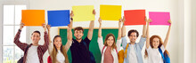 Group Of Happy Diverse School Or College Students Showing Colorful Mockup Banners. Several Smiling Young People Standing In Row And Holding Up Blank Orange, Blue, Yellow, Red And Pink Sheets Of Paper
