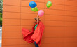 © VISTA by Westend61 - Woman wearing cape holding multi colored balloons in front of wall