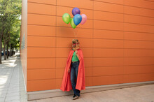 Young Woman Wearing Crocodile Mask And Cape Standing With Multi Colored Balloons In Front Of Wall
