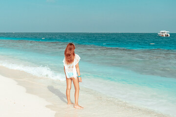 red haired cute teenage girl in swimsuit and cape stands on shore of the Indian Ocean in Maldives island, summer vacation