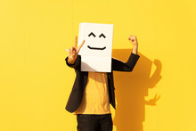Man Wearing Box With Smiley Face Gesturing Peace Sign In Front Of Yellow Wall