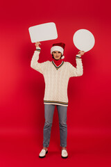 Wall Mural - full length of young woman in santa hat and sweater holding speech bubbles on red.