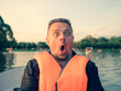 Middle aged handsome man wearing nautical lifejacket afraid and shocked with surprise expression, fear and excited face on lake background.