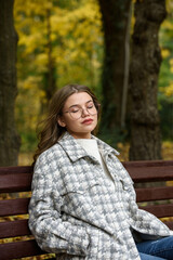 Sticker - European elegant young woman in a stylish trench coat in a white sweater and blue jeans resting on the bench. warm autumn day.