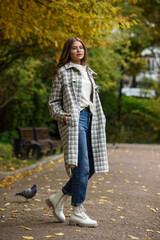 Poster - European elegant young woman in a stylish trench coat in a white sweater and blue jeans walking in a park. warm autumn day.