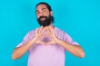 © Jihan - Serious young bearded man wearing violet T-shirt over blue studio background keeps hands crossed stands in thoughtful pose concentrated somewhere