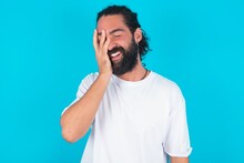 Young Bearded Man Wearing White T-shirt Over Blue Studio Background Makes Face Palm And Smiles Broadly, Giggles Positively Hears Funny Joke Poses